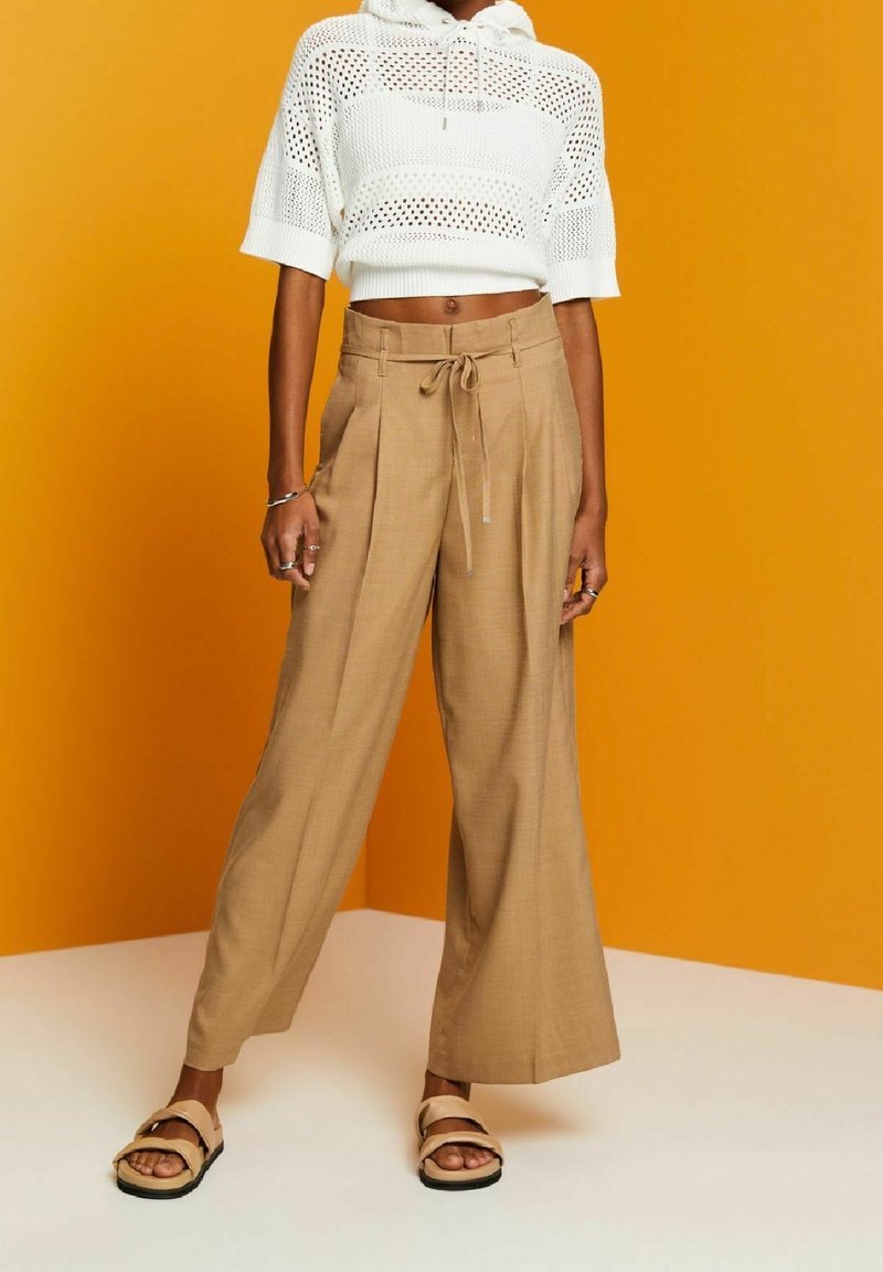 Beige wide-leg trousers with front pleats and drawstring waist. Worn with a cropped white open-knit top. Background is a solid orange.