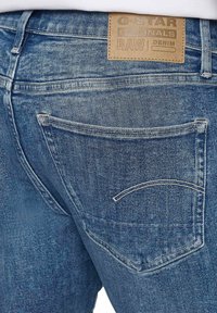 I jeans in denim blu presentano una vista posteriore con una scoloritura più chiara, due tasche posteriori e un'etichetta in pelle beige con dettagli del marchio impressi.
