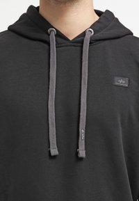 Sudadera negra con capucha y diseño de escote en V, cordones grises y una pequeña etiqueta con el nombre de la marca. Textura suave, ajuste casual.