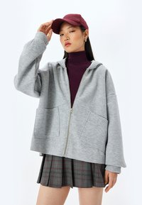 Grå hættejakke med lynlås og rummelig pasform, store frontlommer, parret med en lilla bluse med høj hals og ternet nederdel; modelen bærer en maroon cap.