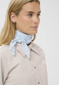 Bufanda de patrón azul claro atada al cuello. Hecha de un tejido suave y ligero. Usada con una camisa beige con cuello que presenta un bolsillo.