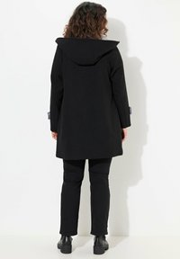 Cappotto di lana nero con un grande cappuccio, caratterizzato da un design strutturato, maniche lunghe e un taglio dritto. Abbinato a pantaloni neri e stivali alla caviglia.