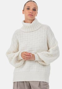 Kuschliger, weißer Strickpullover mit hohem, geripptem Kragen, der ein strukturiertes Korbmuster und ballonförmige Ärmel aufweist. Weiches Material.