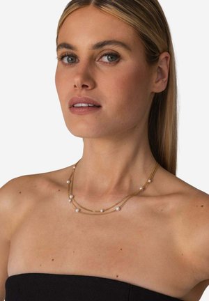 Blonde Frau mit blauen Augen trägt eine doppellagige goldene Halskette mit gleichmäßig verteilten weißen Perlen, bekleidet mit einem schwarzen trägerlosen Oberteil.