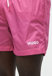 Φωτεινά ροζ μαγιό σορτς με λευκό κορδόνι, φορεμένα από άτομο με σκούρο δέρμα, με λευκό λογότυπο "HUGO" στο αριστερό πόδι.