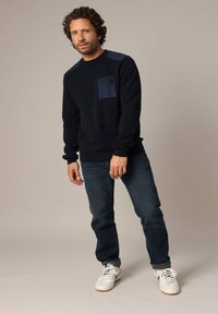 Pull marin avec une poche texturée, des manches longues et des poignets côtelés, associé à un jean bleu foncé et des baskets blanches.