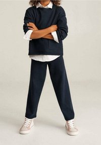 Sudadera azul marino con un bolsillo frontal, combinada con pantalones a juego. Camisa blanca con cuello debajo. Zapatillas de color claro con cordones.