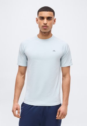 Giovane uomo con capelli corti che indossa una maglietta sportiva a maniche corte azzurro chiaro e pantaloni blu scuro, in piedi davanti a uno sfondo bianco semplice.