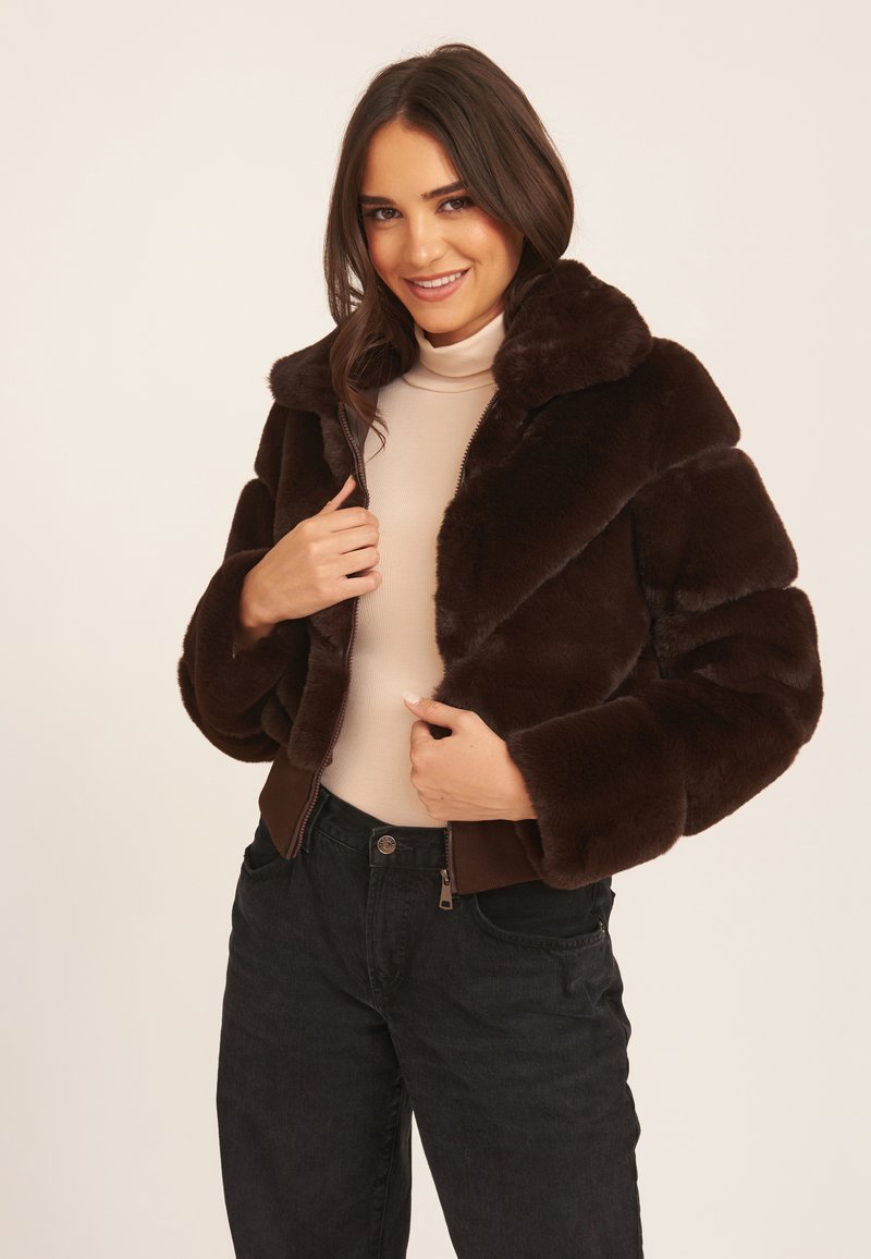 Brown faux fur cropped jacket with large collar, zip closure, and ribbed hem. Worn over a beige top with black jeans.