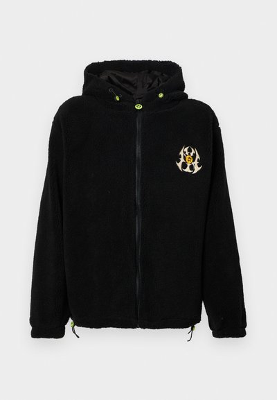 Fekete polár kapucnis pulcsi cipzárral, mellkason logóval, zöld húzózsinórral és állítható alsó részhez tartozó füllel.