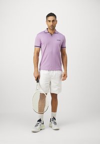 Violet polo marškiniai su kontrastinga juoda apykakle ir rankovių juostomis, derinami su baltais šortais, laikant teniso raketę, dėvint baltus sportinius batus.