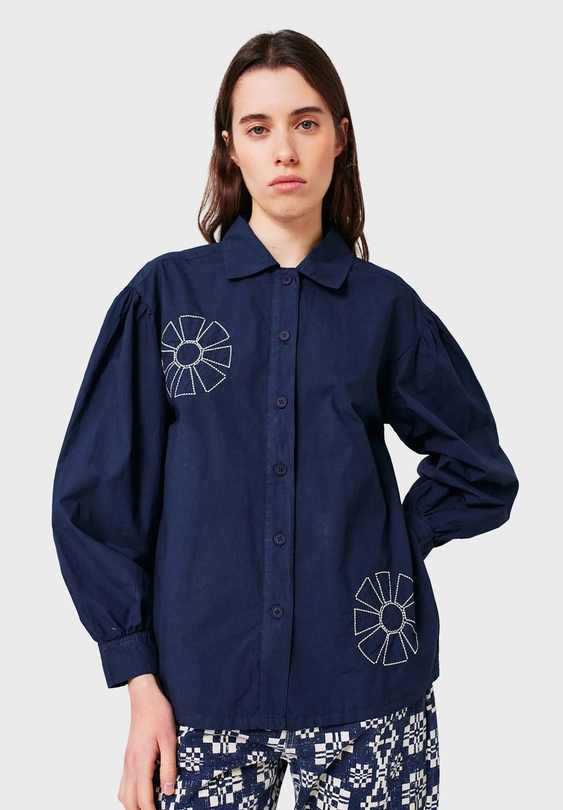 Donna che indossa una camicia button-up blu navy con maniche a sbuffo e disegni floreali ricamati bianchi, abbinata a pantaloni a motivi blu navy e bianchi.