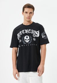Schwarzes Grafik-T-Shirt mit Schädel- und Blumendesigns, großen Schriftzügen und kurzen Ärmeln. Aus Baumwolle mit einem lockeren Schnitt.
