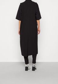 Dlouhý černý kabát bez rukávů se střihem do rovna, doplněný černými kalhotami a černobílými botami na podpatku se vzorem zebry.