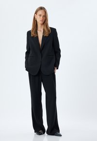 Svart skräddarsydd blazer med en djup V-ringning, kompletterad med matchande vida byxor. Slät textur, minimalistisk design och inga synliga mönster.