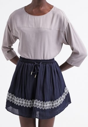 Osoba ubrana w jasno beżową bluzkę z długim rękawem i granatową spódnicę z białymi haftowanymi wzorami oraz troczkiem w pasie.