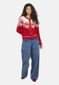 Cardigan rosso in maglia con spalle bianche a motivo, caratterizzato da una trama a treccia, scollo rotondo e bottoni effetto legno; abbinato a jeans a gamba larga blu.