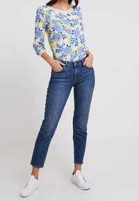 Vrouw in een blauw en geel bloemen blouse met lange mouwen, hoogtaille blauwe jeans en witte sneakers, staande met haar handen in de zakken.