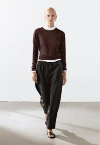 Un pull cropped marron foncé sur une chemise blanche, associé à un pantalon de jogging noir et des sandales plates à lanières, sur un fond de sol neutre.