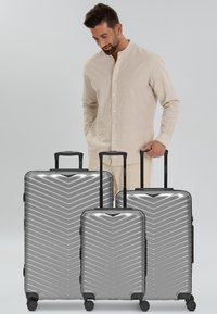 Homme en chemise beige se tient derrière trois valises rigides argentées avec des poignées extensibles sur un fond gris clair.
