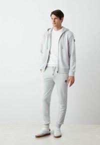 Gaiši pelēka kapucīte ar rāvējslēdzēju un auklu, kombinēta ar atbilstošām sporta biksēm. Tām ir elastīgas aproces un maza logo uz biksēm. Baltas kedas.