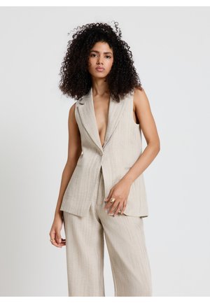 Frau mit lockigem Haar, die einen ärmellosen beigen Blazer und passende weite Hosen trägt, steht vor einem schlichten hellen Hintergrund.