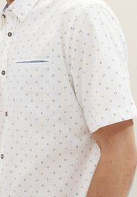 Camicia a maniche corte in tessuto leggero con sfondo bianco, caratterizzata da un motivo geometrico in nero e arancione, e una tasca sul petto.