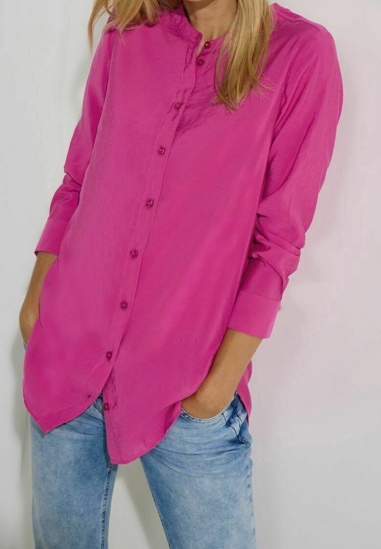 Femme portant une chemise rose vif à manches longues boutonnée, les mains dans les poches d'un jean bleu clair, devant un fond uni.