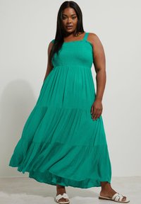 Teal Maxi-Kleid mit verstellbaren Trägern, gerafftem Oberteil und gestuftem Rock. Leichte Stoffqualität mit fließender Textur. Flache weiße Sandalen.