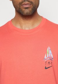 Coral cotton t-shirt featuring a round neckline, short sleeves, and a graphic logo on the left chest in multicolored tones and black accents.