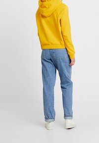 Persona che indossa un vivace felpa gialla, jeans azzurri arrotolati e sneakers bianche, in piedi contro uno sfondo bianco uniforme, con la schiena rivolta verso l'osservatore.
