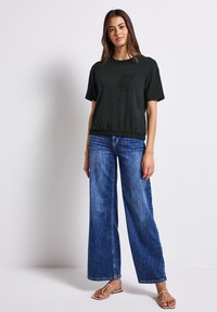 Černé tílko s krátkým rukávem, kapsou a elastickým lemem, spárované s širokými modrými džínami a hnědými sandály. Hladký materiál, ležérní design.