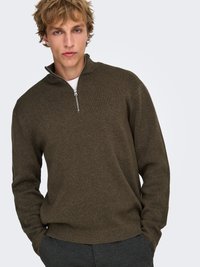 Pull marron à fermeture éclair en tissu tricoté texturé avec un col montant et des manches longues, associé à un pantalon gris foncé.
