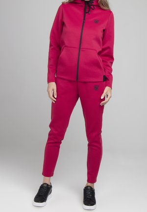 Femme portant un sweat à capuche rouge zippé et un pantalon de survêtement assorti avec des baskets noires, debout devant un fond gris uni.