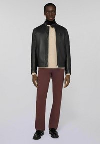 Schwarze Lederjacke mit hohem Kragen, getragen über einem hellbeigen Pullover und kastanienbraunen Hosen. Das Model steht mit den Händen an den Seiten und trägt schwarze Schuhe.