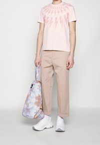 Světle růžové tričko s designem blesku, béžové croppované kalhoty, bílé tenisky a batoh s pastelovým tie-dye vzorem na stažení.