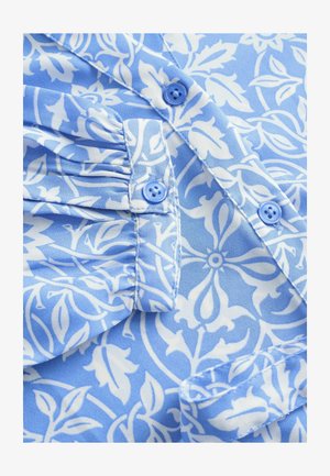 Blåt blomstermønstrede stof har en lys blå baggrund med hvide designs, fremhævet af blå knapper og samlede detaljer på skulderstykkerne.