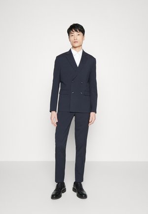 Costume bleu marine croisé avec une coupe ajustée, présentant un revers cranté et deux poches avant, associé à des chaussures de ville noires.