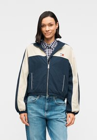 Μπλε ναυτικό και κρεμ μπουφάν με φερμουάρ, όρθιο γιακά, ελαστικά μανίκια και λογότυπο σε εμφανές σημείο στο στήθος. Συνδυάζεται με ανοιχτόχρωμο μπλε τζιν.