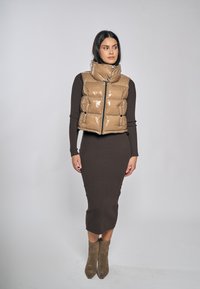 Prošívaná vesta v lesklé barvě taupe s vysokým límcem, zapínáním na zip a dvěma předními kapsami, nošená přes přiléhavé hnědé žebrované šaty a kotníkové boty.