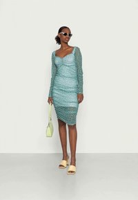 Vestido justo de manga longa, na cor verde-água, com bolinhas brancas; textura franzida; combinado com sapatos amarelos e uma pequena mala verde.