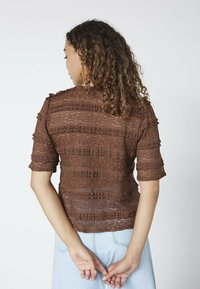 Kvinde med krøllet hår iført en brun blonde-top med flæser og lyseblå jeans, stående med hænderne foldet bag ryggen.