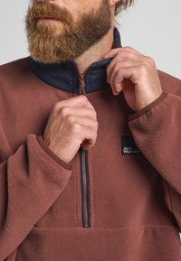 Pull en polaire de couleur brun rouillé avec un col marine, fermeture zippée à moitié et patch logo ; texture douce avec des poignets et un ourlet inférieur côtelés.