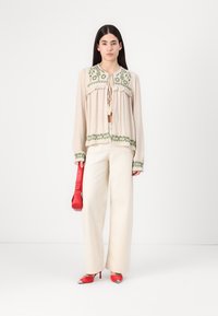 Blouse beige avec broderies florales vertes, fermeture à pompon, associée à un pantalon large ivoire et des escarpins rouges à bout pointu, tenant un sac rouge.