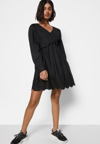 Vestido de algodão preto com mangas longas, decote em V, detalhe de babados no peito e bainha de renda bordada. Usado com ténis deportivos pretos.