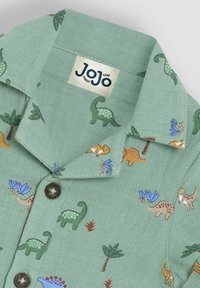 Grünes kurzärmliges Hemd mit Dinosauriermotiv, braunen Knöpfen und Etikett. Der Stoff hat eine strukturierte, gewebte Optik.