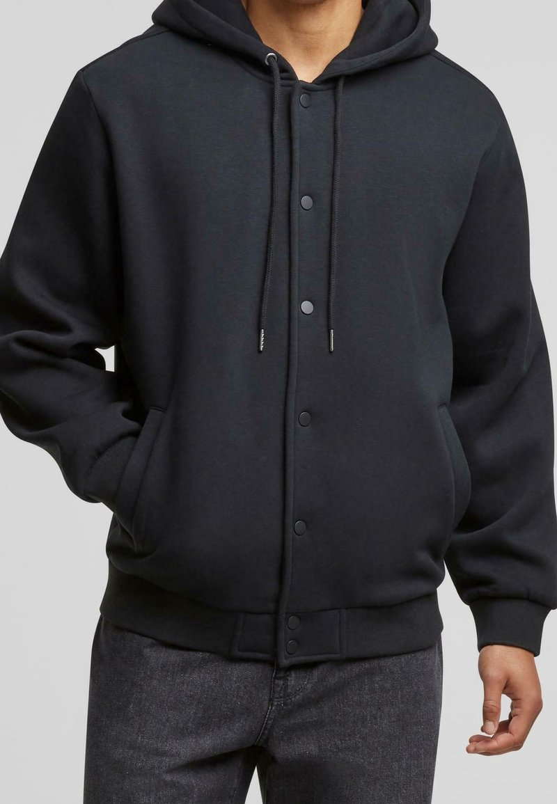Homme portant une veste noire à capuche avec boutons-pression, cordons, poches latérales et poignets côtelés, associée à un jean en denim foncé.