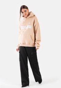 Beżowa bluza z kapturem z białym haftem, dużą kieszenią i sznurkiem do regulacji kaptura, w połączeniu z luźnymi czarnymi spodniami cargo z wieloma kieszeniami.