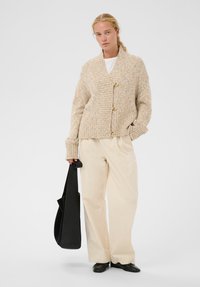 Frau trägt eine beigen, grob gestrickten Cardigan, cremefarbene, weit geschnittene Hosen, schwarze Schuhe und hält eine große schwarze Tasche, steht vor einem einfarbigen Hintergrund.