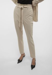 Beige op maat gemaakte broeken met een hoge taille en strikceintuur, met zijzakken en een gladde textuur. Gecombineerd met zwarte puntige hakken.